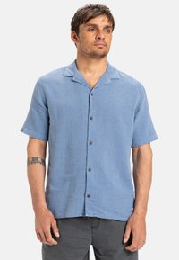 Svetlo modrá krátkopp rukávová košeľa na gombíky vyrobená z tkaniny s prešívaním, s vykrojeným golierom, hnedými gombíkmi a voľným strihom.