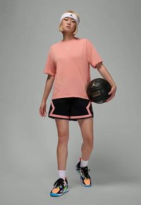 Naine kannab roosat korvpalli särki ja musti lühikesi pükse, hoides musta korvpalli, seistes halli tausta ees.