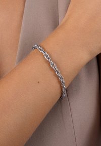 Bracelet en chaîne en métal argenté avec des maillons entrelacés, présentant un design torsadé. Le poignet apparaît sur un fond en tissu neutre.