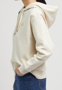 Sudadera beige con bolsillo canguro, capucha con cordón, puños y dobladillo acanalados, que presenta un discreto bordado del logo en el frontal.