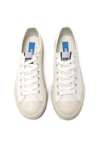 Zapatillas de lona blancas con suelas de goma de color claro, con punta redondeada, cierre de cordones y una etiqueta de marca en la lengüeta en azul y negro.