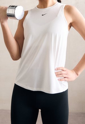 Woman in white Nike tank top lifts silver dumbbell with one arm, other hand on hip, indoors against plain background.