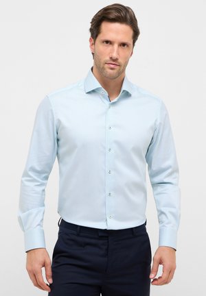 Mand iført lyseblå tekstureret skjorte med knapper og mørke bukser, stående mod en ensfarvet lys baggrund.