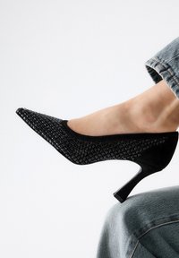 Zapato negro de tacón alto con punta afilada y parte superior texturizada y trenzada. Presenta un tacón delgado y detalles suaves en un diseño clásico.