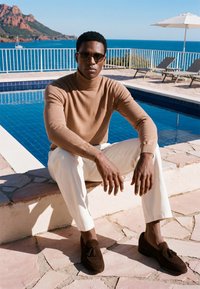 Homme portant des lunettes de soleil, un col roulé marron, un pantalon blanc et des mocassins marron, assis près d'une piscine avec l'océan et les montagnes en arrière-plan.