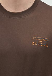 Καφέ βαμβακερό μπλουζάκι με ριμπ λαιμόκοψη. Στο μπροστινό μέρος υπάρχει πορτοκαλί γραφικό κείμενο που αναγράφει "PROPERTY OF DIESEL" με επικάλυψη σχεδίου.