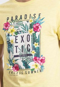 T-shirt en coton jaune présentant un design floral vibrant avec des fleurs d'hibiscus roses et jaunes, des feuilles tropicales, et le texte "VACANCES EXOTIQUES."