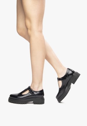 Piernas con zapatos de tiras en T negros brillantes con suela gruesa y textura de cocodrilo, posando a medio paso sobre un fondo blanco.