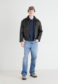 Veste en cuir synthétique noire avec fermeture éclair, chemise bleu foncé, jeans bleu clair, chaussures grises et casquette marron avec un logo.