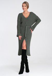 Olivgrünes, geripptes Strickkleid mit V-Ausschnitt, drei Knöpfen, seitlichem Schlitz und tailliertem Gürtel. Das Model trägt schwarze kniehohe Stiefel.