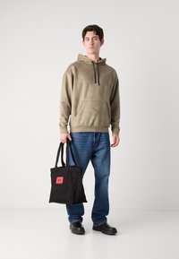 Hoodie cáqui com um bolso frontal, jeans azuis e uma bolsa tote preta com um rótulo vermelho. Modelo usa sapatilhas pretas.
