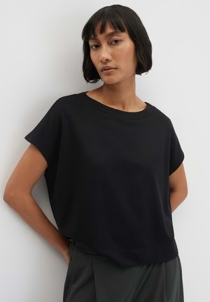 Vrouw met kort donker haar draagt een losse zwarte top met korte mouwen en donkere broek, staand tegen een witte muur.