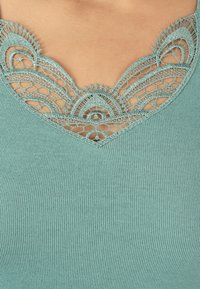 Top de color verde claro con escote en V, con intrincados detalles de encaje y textura acanalada en el cuerpo. El encaje presenta un patrón floral con bordes ondulados.