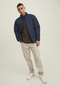 Giacca in nylon trapuntata blu marina con colletto alto, abbinata a una camicia scura e pantaloni beige chiaro. Sneakers bianche completano l'outfit.