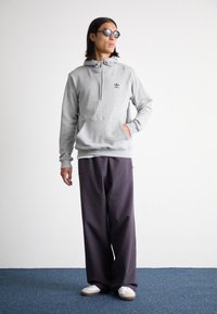 Felpa grigia con cappuccio, tasca frontale e logo nero, abbinata a pantaloni larghi viola scuro e sneaker bianche su uno sfondo blu.