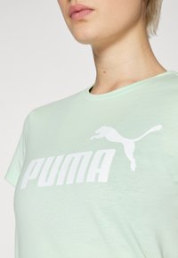Svetlozelené bavlnené tričko s výrazným bielym logom "PUMA" a grafikou skáčuceho pumy. S okrúhlym výstrihom a krátkymi rukávmi.