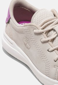 Ljusbeige sneaker med en texturerad mockaöverdel, meshdetaljer, lila akcentfärger på hälen och en vit gummisula. Präglat logo finns.