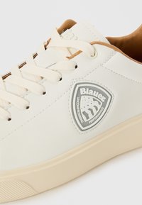 Adidași sintetici albi cu talpă din cauciuc bej, design cu șireturi și un logo triungiular argintiu care include text și detalii grafice.
