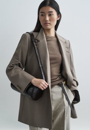 Blazer beige surdimensionné, haut en tricot marron, pantalons gris et sac à main texturé foncé. Détails notables : grandes manches, accents dorés sur la ceinture, silhouette structurée.