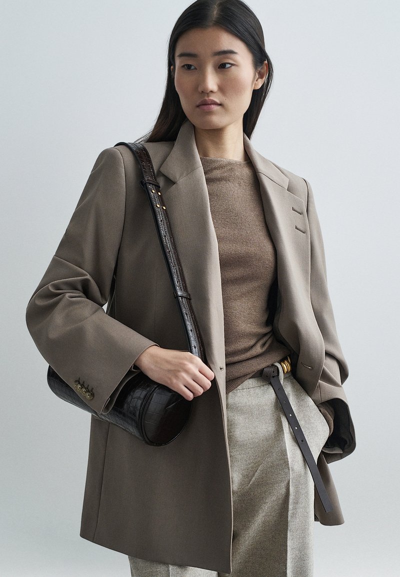 Μπεζ υπερμεγέθη σακάκι, καφέ πλεκτό τοπ, γκρι παν�τελόνι και σκούρα υφασμάτινη τσάντα. Σημαντικές λεπτομέρειες: μεγάλα μανίκια, χρυσές λεπτομέρειες στην ζώνη, δομημένη σιλουέτα.