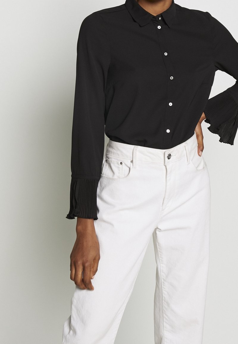 Blusa nera a maniche lunghe con colletto, chiusura a bottoni e dettagli plissettati sui polsini, abbinata a jeans bianchi a vita alta.