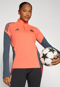 Bright coral orange and dark gray long-sleeve athletic shirt with a high collar and Adidas stripes. Model holds a black-and-white soccer ball.