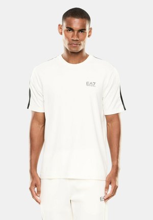 Giovane uomo che indossa abbigliamento sportivo bianco EA7 Emporio Armani, in piedi davanti a uno sfondo chiaro e uniforme, rivolto in avanti.