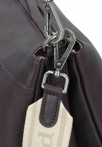 Detalle de un bolso de cuero morado con herrajes plateados, un clip y costuras en contraste. Textura suave con una correa texturizada y etiqueta de logo.
