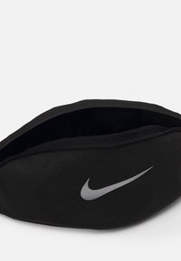 Svart Nike-väska med en strukturerad yta, dragkedjestängning och en silverfärgad swoosh-logga på ena sidan. Formen är oval och strömlinjeformad.