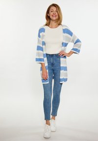 Gestreifter Strickcardigan in Blau und Weiß, offene Vorderseite mit Ärmeln bis zum Ellbogen. Trägt man über einem cremefarbenen Oberteil und kombiniert mit blauen Jeans und weißen Sneakers.