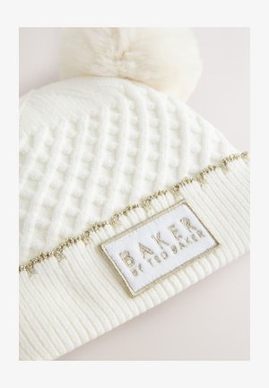 Bonnet en maille blanche présentant un motif texturé, un pompon en fausse fourrure douce et une bande dorée avec un patch logo.
