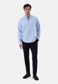 Camicia azzurra chiara con colletto alla cinese e tre bottoni, abbinata a pantaloni scuri su misura e scarpe eleganti nere senza lacci.