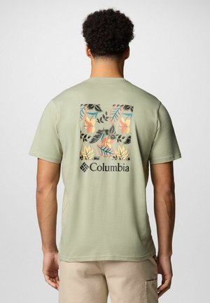 Mann trägt hellgrünes Columbia-T-Shirt mit buntem Blumen- und Blättermuster und Logo auf dem Rücken, beige Hose von hinten gezeigt.