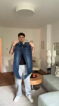 Ξεθωριασμένα μπλε τζιν παντελόνια υψωμένα, εμφανίζοντας ένα τυπικό σχέδιο πέντε τσεπών και κλείσιμο με κουμπί, εκτεθειμένα σε ένα φωτεινό δωμάτιο με απλά έπιπλα.