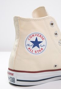 Baskets montantes en toile crème avec œillets en métal, accents rouges et bleus, arborant un logo circulaire avec une étoile bleue et du texte.