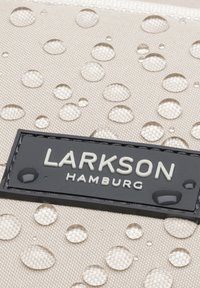 Beige szövet pénztárca texturált felülettel, vízcseppekkel, és egy fekete logós címkével, amelyen az olvasható: "LARKSON HAMBURG."