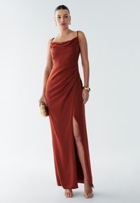 Lång klänning i rostfärgat, mjukt tyg med draperad dropphalsringning och sidospalt. Kombinerad med en guldfärgad clutch och remklackar.