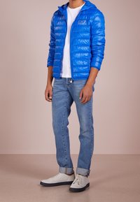 Niebieska kurtka puchowa z kapturem i pikowaną teksturą, w zestawie z jasnoniebieskimi dżinsami i białymi trampkami z ciemną podeszwą.