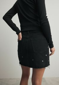 Jupe en denim noir avec ourlet effiloché, accents à œillets argentés et deux poches arrière. Portée avec un t-shirt noir à manches longues, vue de dos.