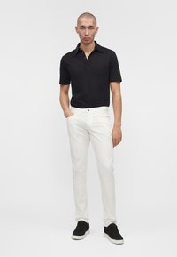 Mann steht mit einer Hand in der Tasche, trägt ein schwarzes kurzärmeliges Poloshirt, weiße Hose und schwarze Sneakers vor einfachem Hintergrund.