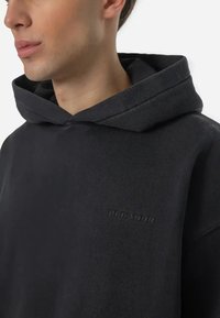 Black hoodie featuring a subtle branded logo in a tone-on-tone design, with a relaxed fit and textured fabric. Close-up view.