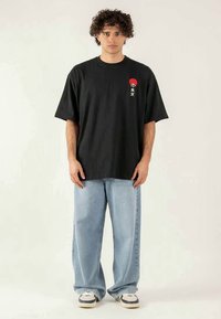 Homem com cabelo encaracolado a usar uma t-shirt preta oversized com gráfico vermelho e jeans largos em azul claro, em pé contra um fundo simples.