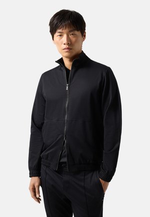 Man wearing a black zip-up jacket with elastic cuffs and black pants, standing with one hand in pocket against plain background.