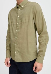 Cămașă button-up de culoare verde măslin, confecționată dintr-un material ușor, având un guler clasic, butoane albe și un mic logo pe piept.