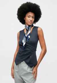 Mulher com cabelo afro, vestindo uma blusa sem mangas azul-marinho, um lenço listrado azul-marinho e branco ao pescoço e calças cinza claro com riscas.