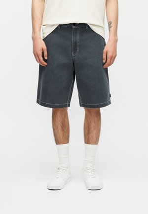 Άνδρας που φοράει σκούρα γκρι σορτς μήκους μέχρι το γόνατο, λευκές κάλτσες και λευκά αθλητικά παπούτσια, στέκεται μπροστά σε ένα απλό φόντο.