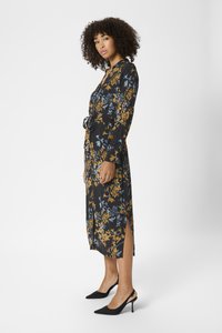 Femeie cu păr creț, îmbrăcată într-o rochie midi neagră cu imprimeu floral galben și albastru, purtând tocuri cu vârf ascuțit în spate, stând de profil.