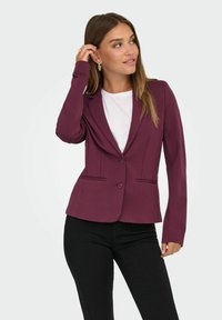 Bordeaux-farvet fitted blazer med to frontlommer, notch-lapel og enkeltknaplukning, båret over en hvid skjorte og sorte bukser.