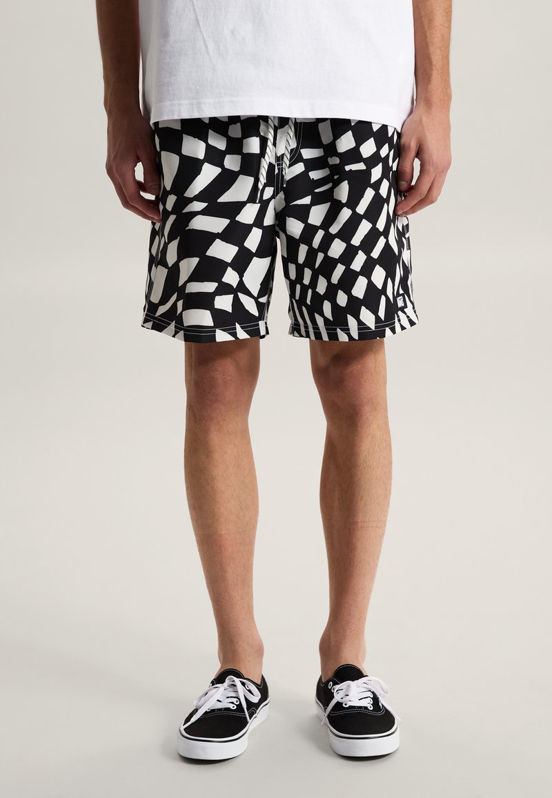 Mand iført sort-hvide shorts med geometrisk mønster, hvid t-shirt og sort-hvide snøresko, stående på en lys baggrund.