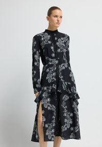 Schwarzes Kleid mit Paisley-Muster, hohem Kragen, langen Ärmeln, mehrlagigem Volant-Saum und Oberschenkelschlitz. Der Stoff wirkt leicht und strukturiert.